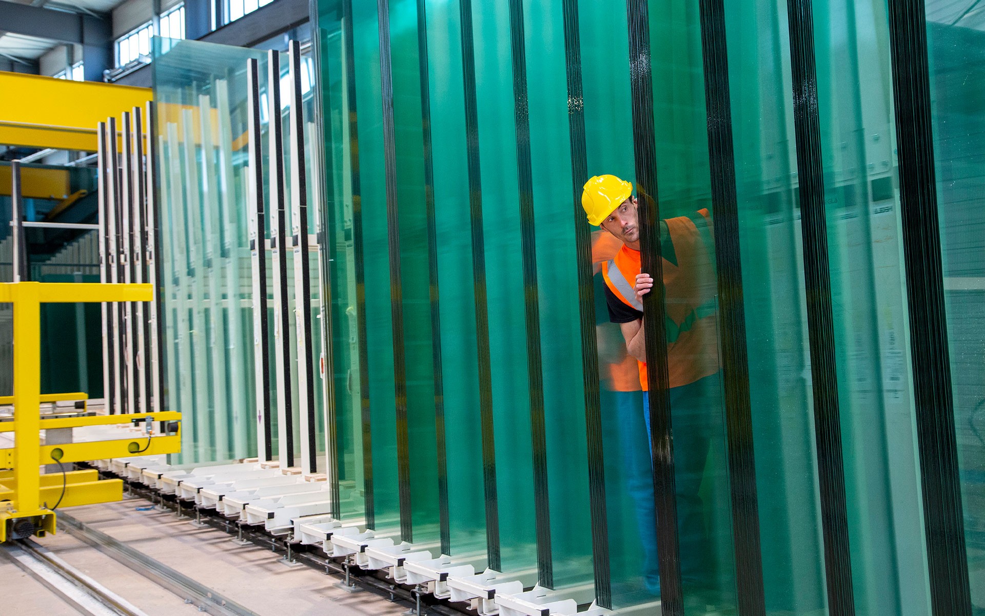 Dans un atelier industriel, un employé portant un casque de chantier jaune et un gilet de sécurité orange est partiellement visible alors qu'il se tient derrière une grande pile de feuilles de verre teinté vert, alignées verticalement sur des supports métalliques. Les plaques de verre créent un effet de répétition visuelle et de transparence, soulignant l'environnement de production industrielle de l'usine.