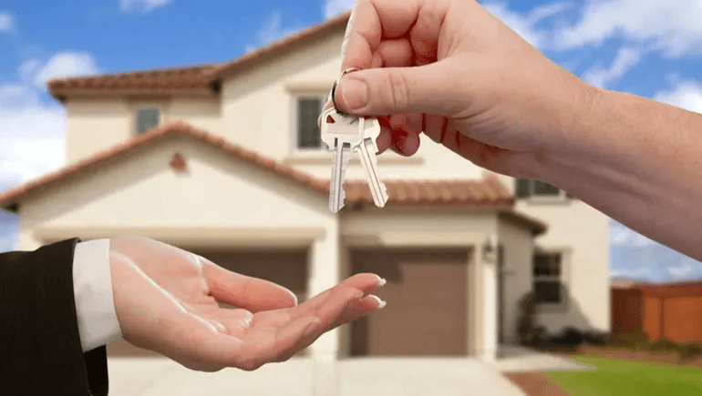 Una mano entrega las llaves de una casa a otra persona frente a una vivienda, símbolo del acceso a la vivienda propia.