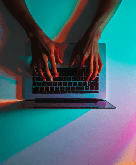 Hands typing on a laptop keyboard, bathed in vibrant teal and pink lighting, creating a dynamic and creative atmosphere.