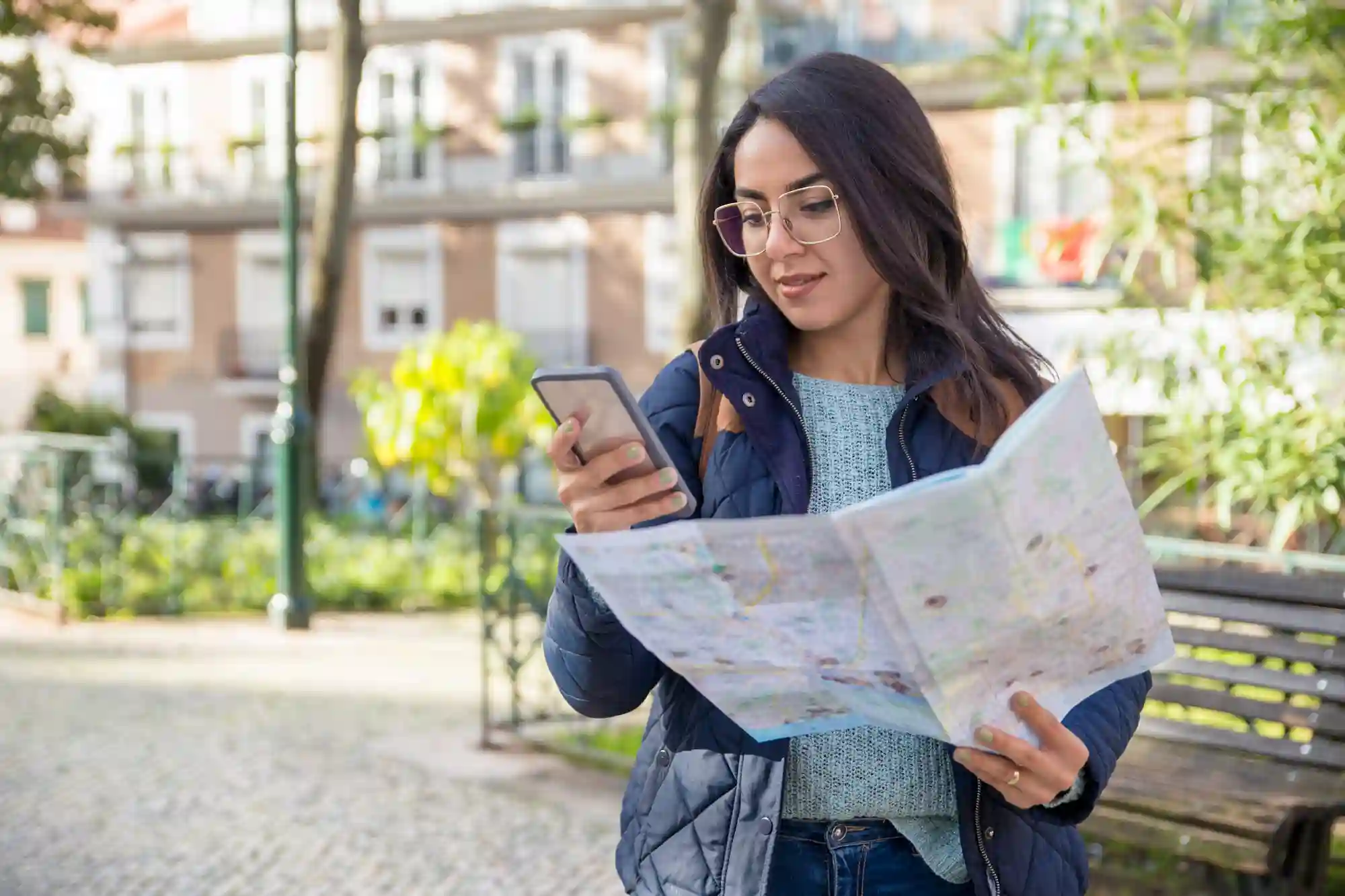 Woman checking her smartphone and a paper map while navigating a city park.