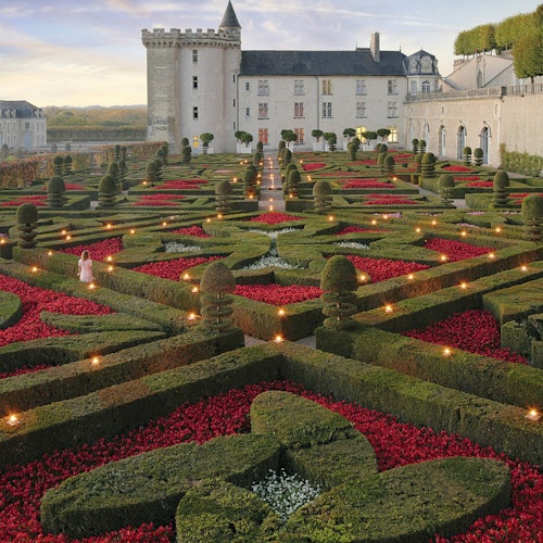 Formal garden with intricate hedges and bright red flowers, leading to a historic castle with a tower and stone walls.