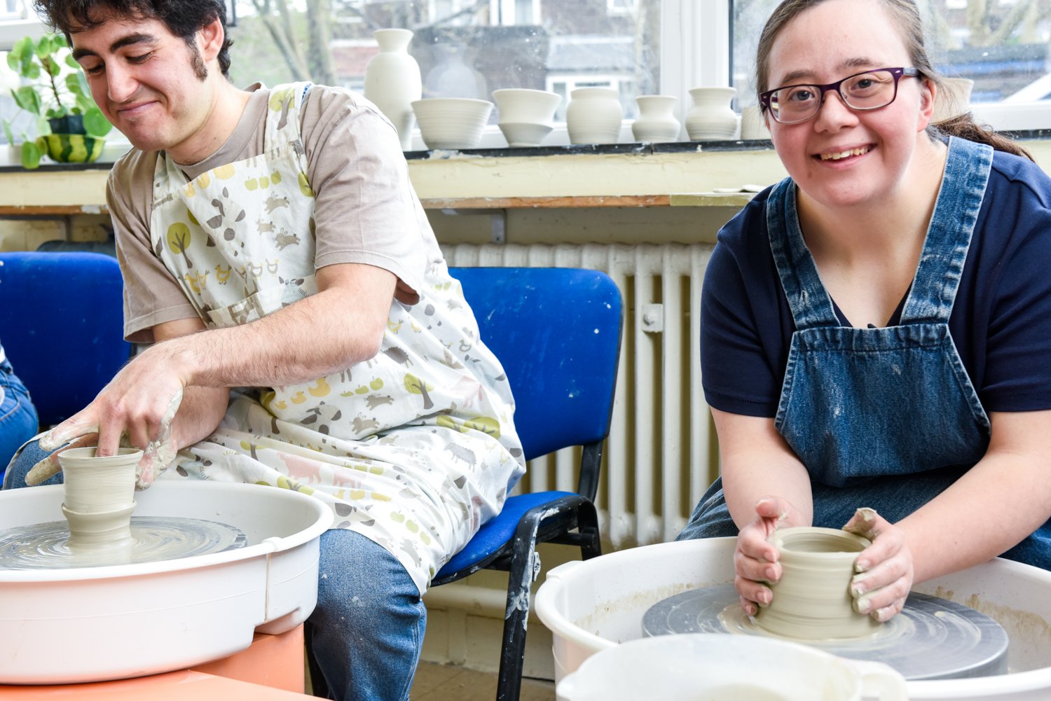 Two people sit at a table covered with art materials, including coloured markers, pens, scissors and glue. Both are working on illustrated paper designs, adding details and colour to architectural-style drawings. The table is filled with creative supplies and works in progress, suggesting a collaborative arts and crafts or design workshop.