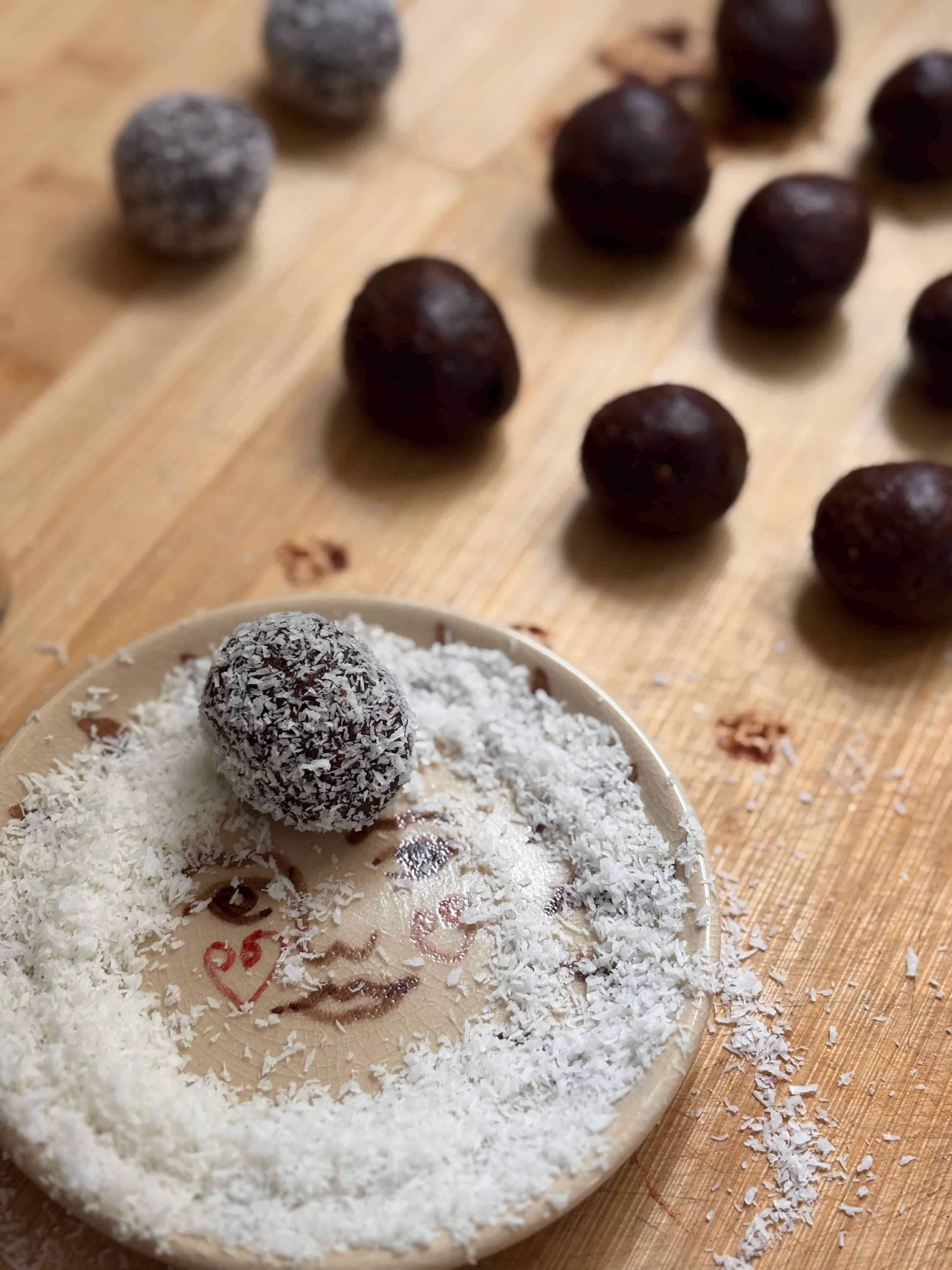 Bliss ball being rolled in shredded coconut on a small plate, with finished chocolate snack balls in the background on a wooden surface.