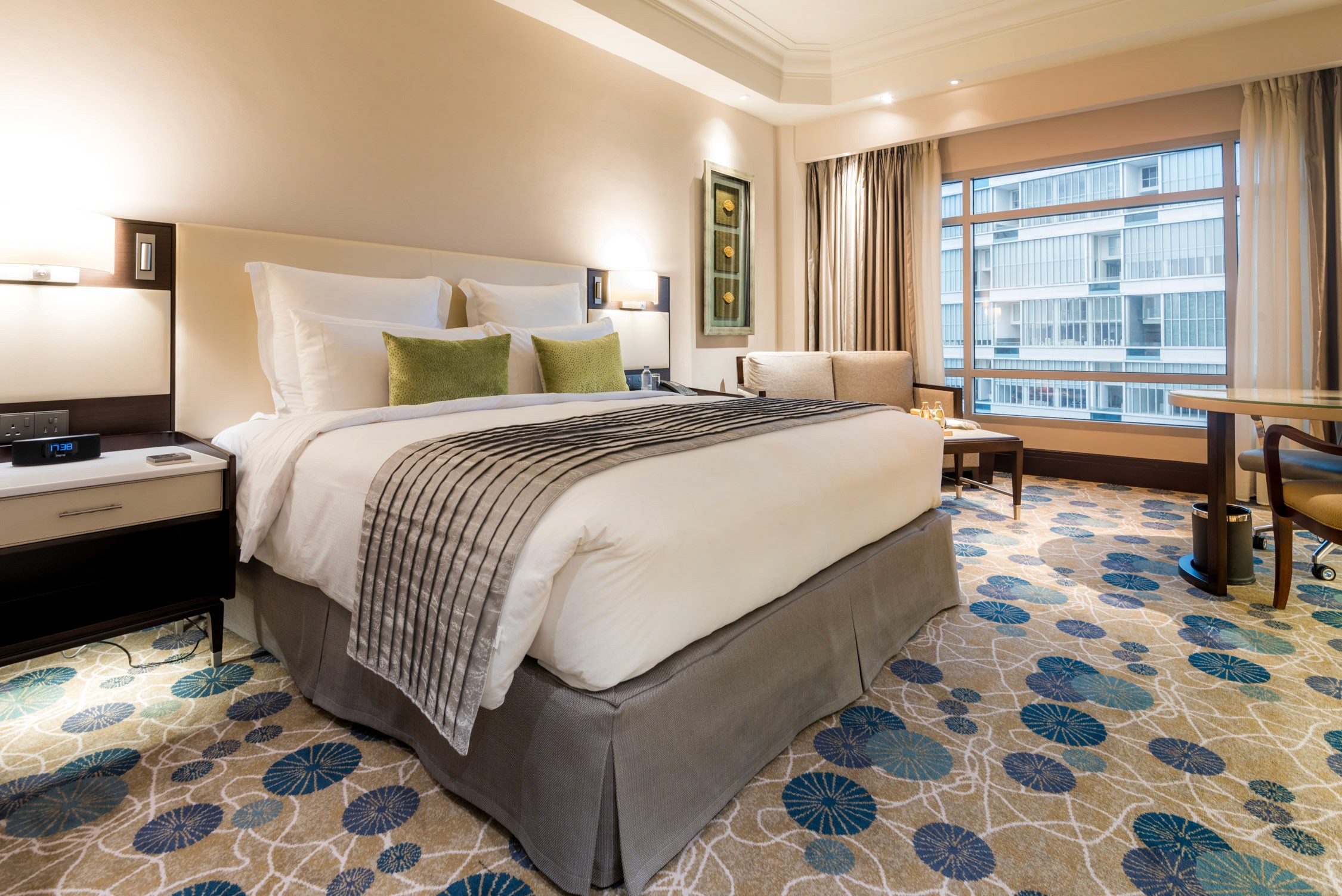 A modern hotel room features a large bed with white linens and green pillows, a patterned carpet, and a window view.