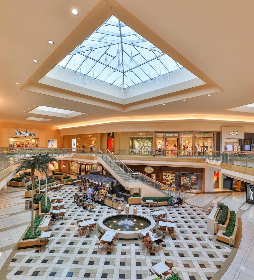 Interior rendering of International Plaza mall showing a shopping corridor with retail storefronts, skylights with natural lighting, decorative geometric flooring, plants, and modern design elements