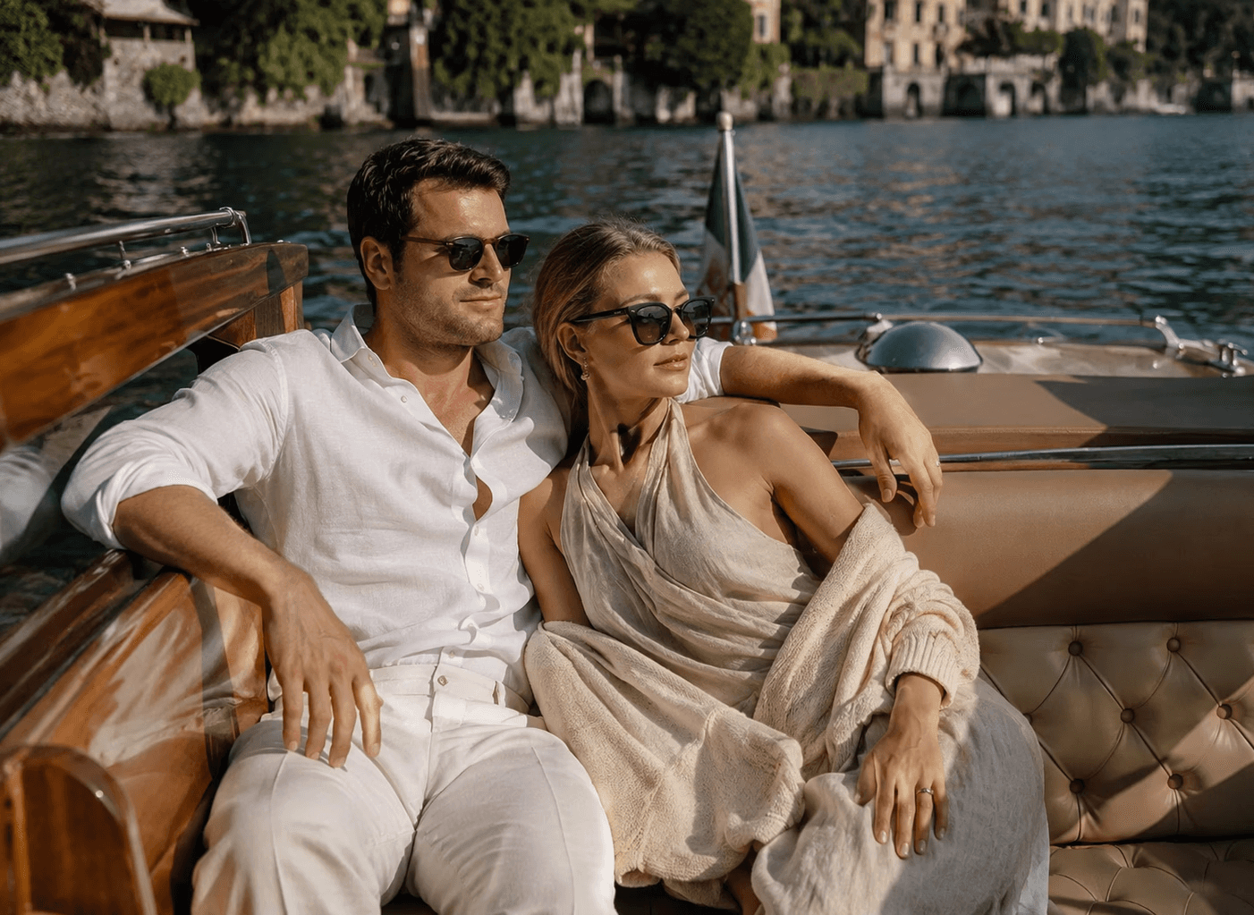 A sunny, medium shot of a stylish couple sitting together on the leather bench of a classic wooden speedboat on Lake Como. The man, wearing dark sunglasses, an unbuttoned white linen shirt, and white trousers, has his arm draped casually around the woman. The woman, also wearing dark sunglasses, wears an elegant cream halter dress with a light cardigan draped over her shoulders. The dark water of the lake and historic coastal villas line the background under bright natural sunlight.