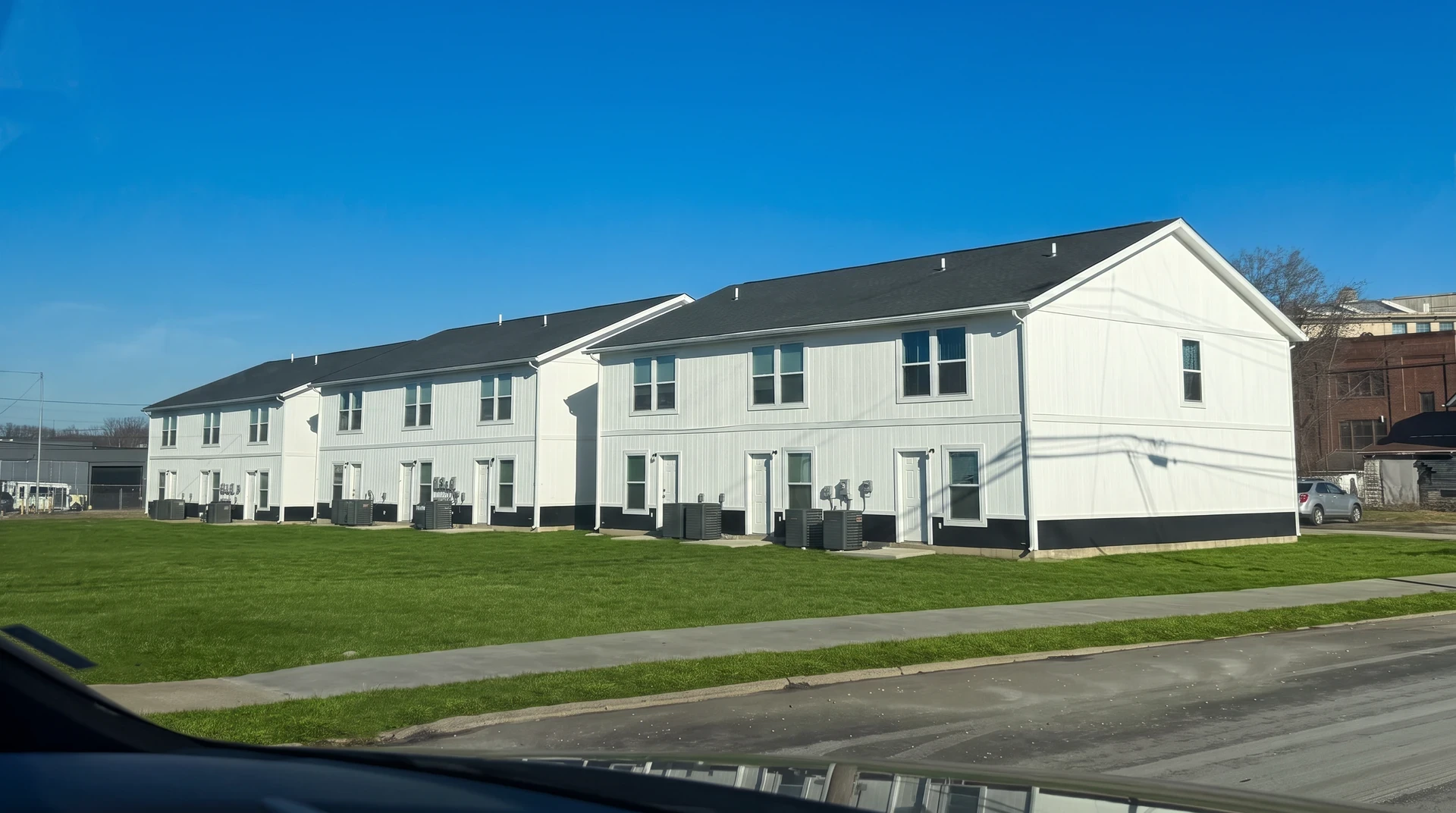 A row of white two-story townhomes with dark grey roofs and skirting, seen across a lawn from the road