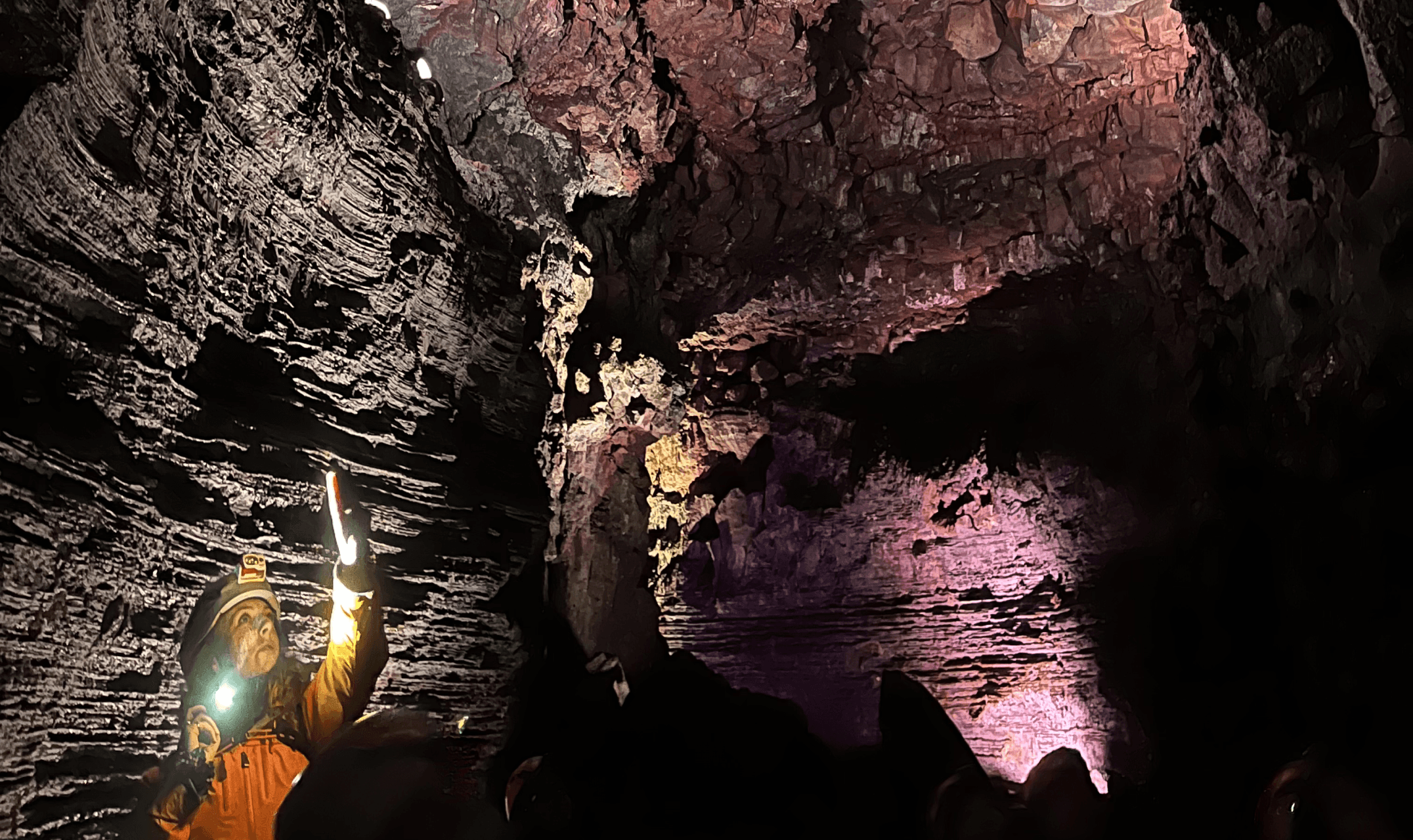 A group explores a lit-up lava cave or magma tunnel with pink and yellow lighting, evoking adventure and team bonding.
