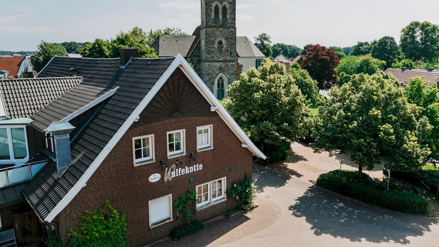 Außenfassade von oben fotografiert, der Gaststätte mit dem Kirchturm im Hintergrund.