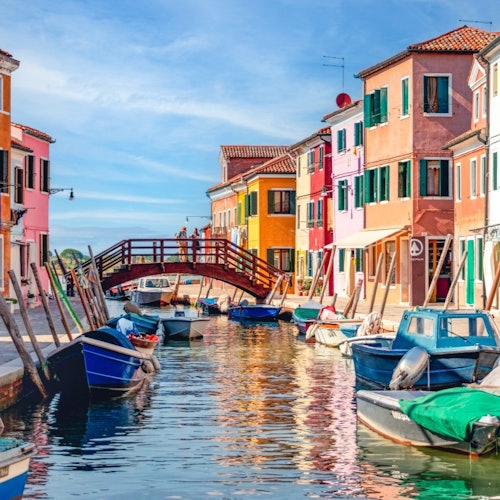 Colorful buildings line a canal with docked boats, under a blue sky. A wooden bridge spans the canal in the background.