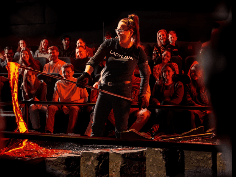 A close-up of two smiling audience members seated at the Lava Show, illuminated by the warm, orange glow of molten lava. Both guests are wearing protective glasses and appear captivated by the spectacle in front of them. The cozy, dark theater setting and the soft lighting create an engaging and intimate atmosphere. One guest holds a drink, adding to the relaxed and enjoyable experience.