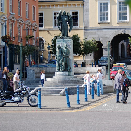 Une place de la ville présente une statue commémorative de guerre en bronze avec des personnes qui se promènent, une moto et des bâtiments colorés en arrière-plan.