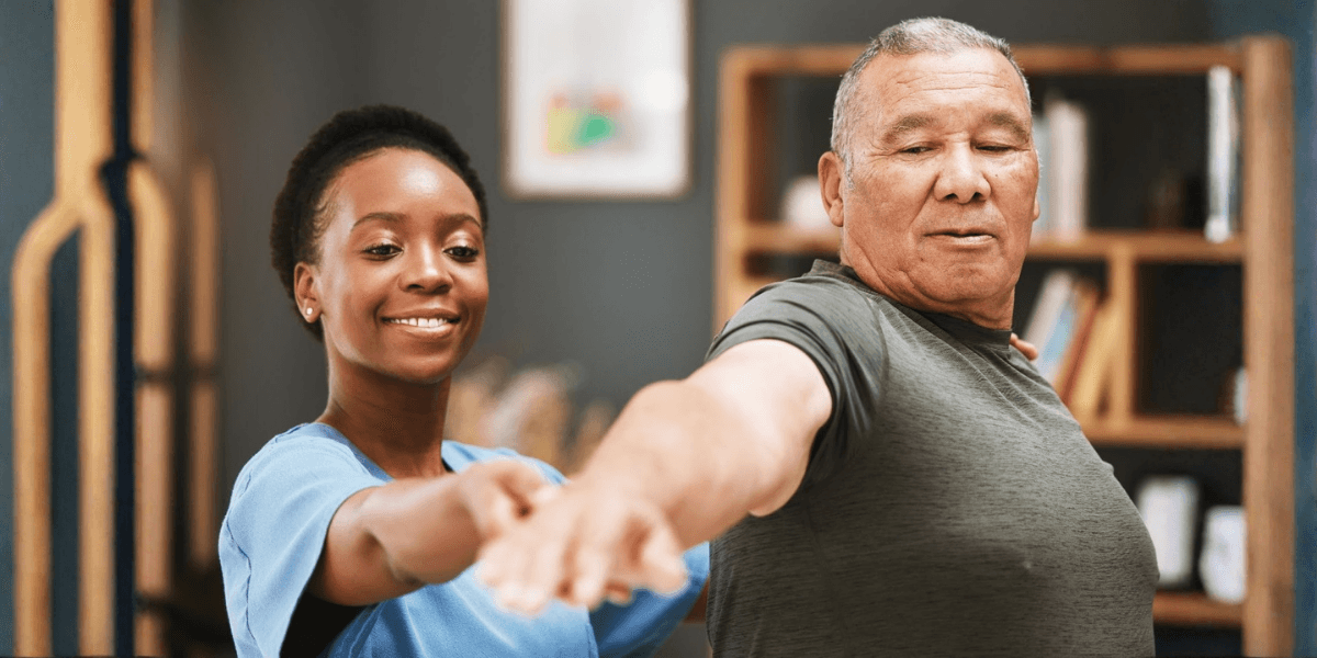 Smiling caregiver assisting an older man with arm stretching in a home care session highlighting trust comfort and personalized assistance.