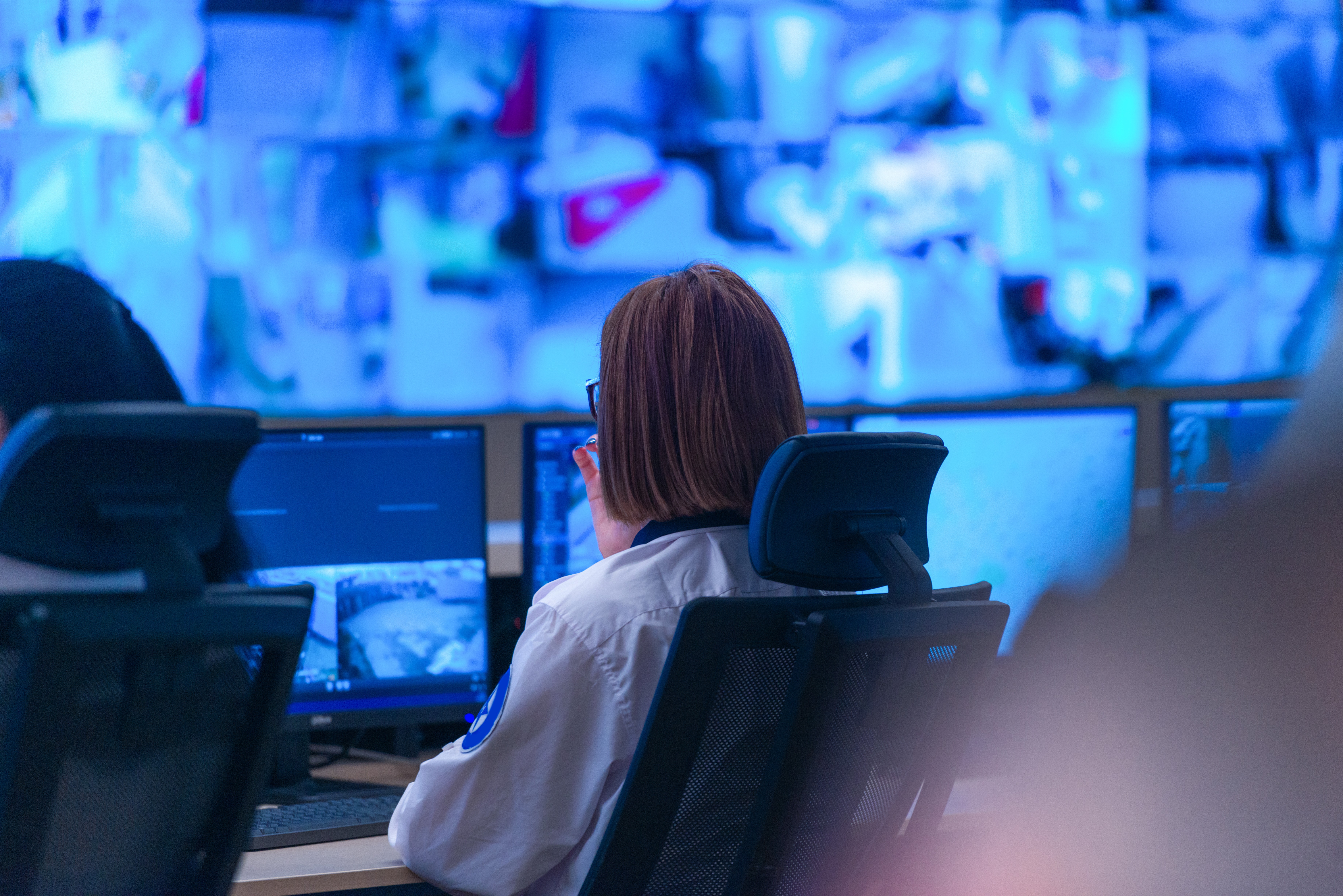Rückenansicht einer Frau in einem hellen Hemd, die in einer Kontroll- oder Überwachungszentrale vor mehreren Monitoren sitzt. Vor ihr auf dem Schreibtisch sind zwei Computerbildschirme zu sehen, die Überwachungsbilder zeigen. Die gesamte Wand hinter ihr besteht aus einer großen Video-Wand, auf der viele kleine Quadrate mit bläulich beleuchteten Überwachungsfeeds laufen. Die Szene ist von einem hellblauen Licht dominiert.