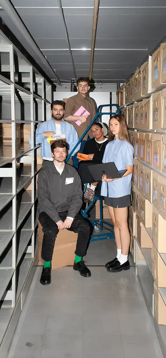 Groupe de cinq étudiants et étudiantes du Collectif1440 posant dans une réserve d’archives, entourés de boîtes en carton et de rayonnages métalliques, chacun tenant des spécimens typographiques.