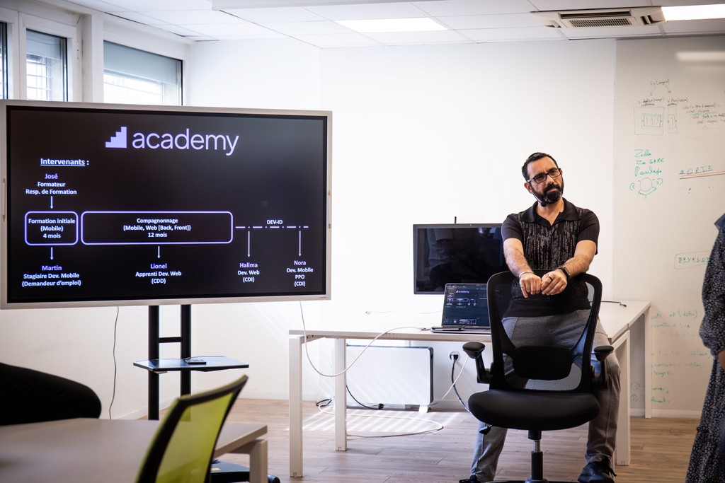 Un homme assis sur une chaise devant une salle de classe, près d’un écran affichant un organigramme décrivant un parcours de formation en développement mobile.