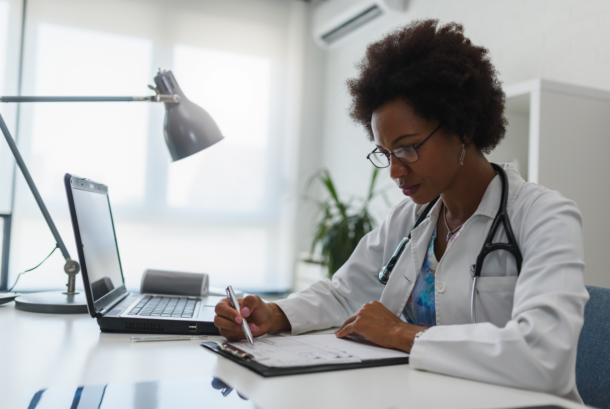 A doctor in a white coat with a stethoscope working on a patient record at her desk, reviewing documents and utilizing a laptop for revenue cycle management tasks.