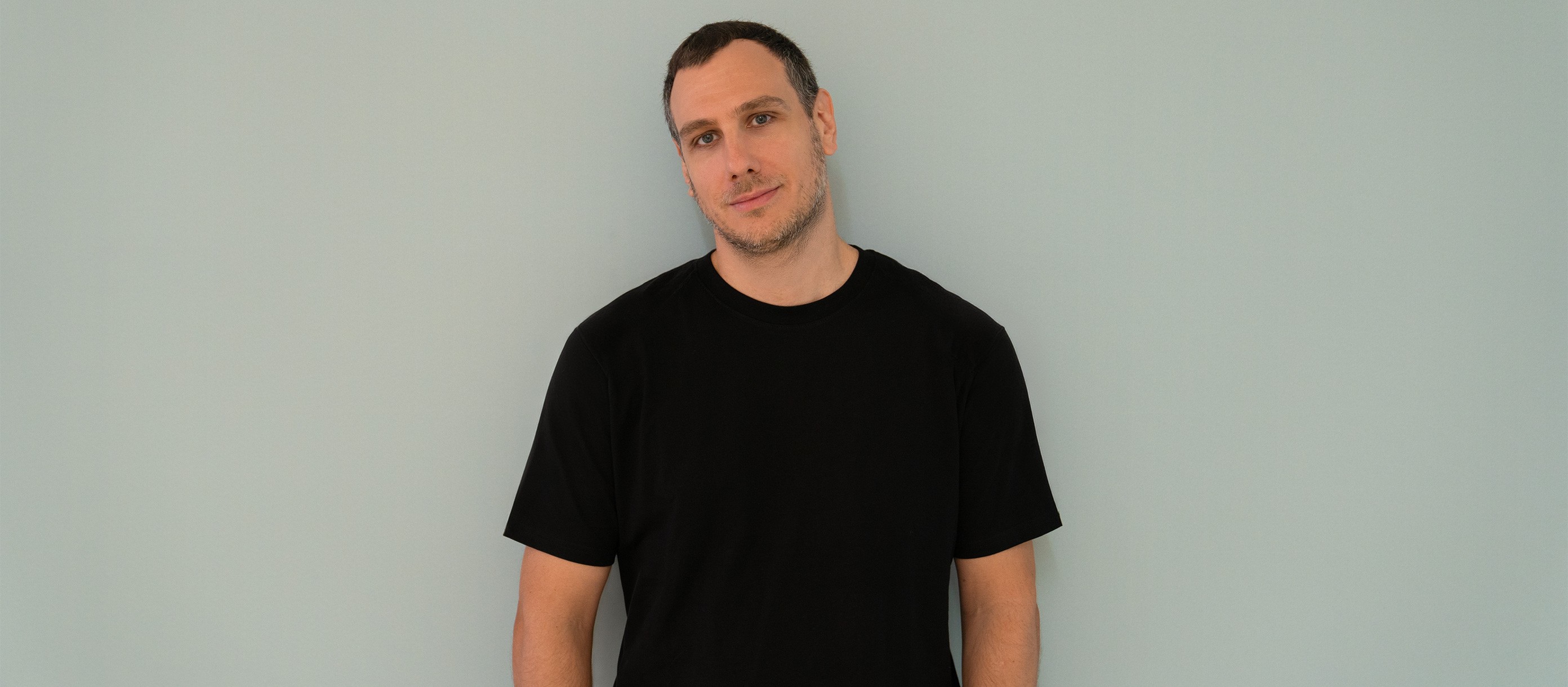 Nik Susic, a creative director, standing against a pale teal wall in a black T-shirt.