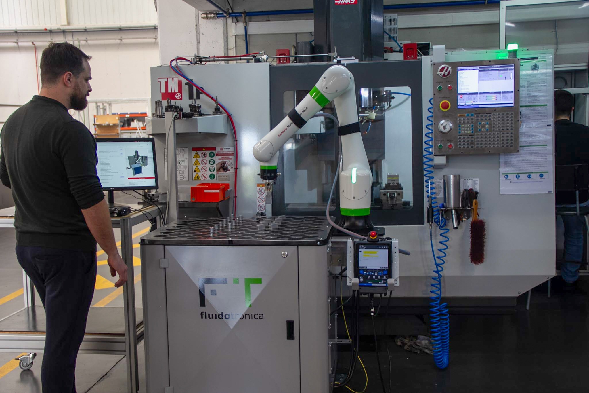 A technician monitoring a robot arm performing machine tending on a CNC mill at Fluidotronica, with the robot's teach pendant and a parts tray staged in front of the machine.