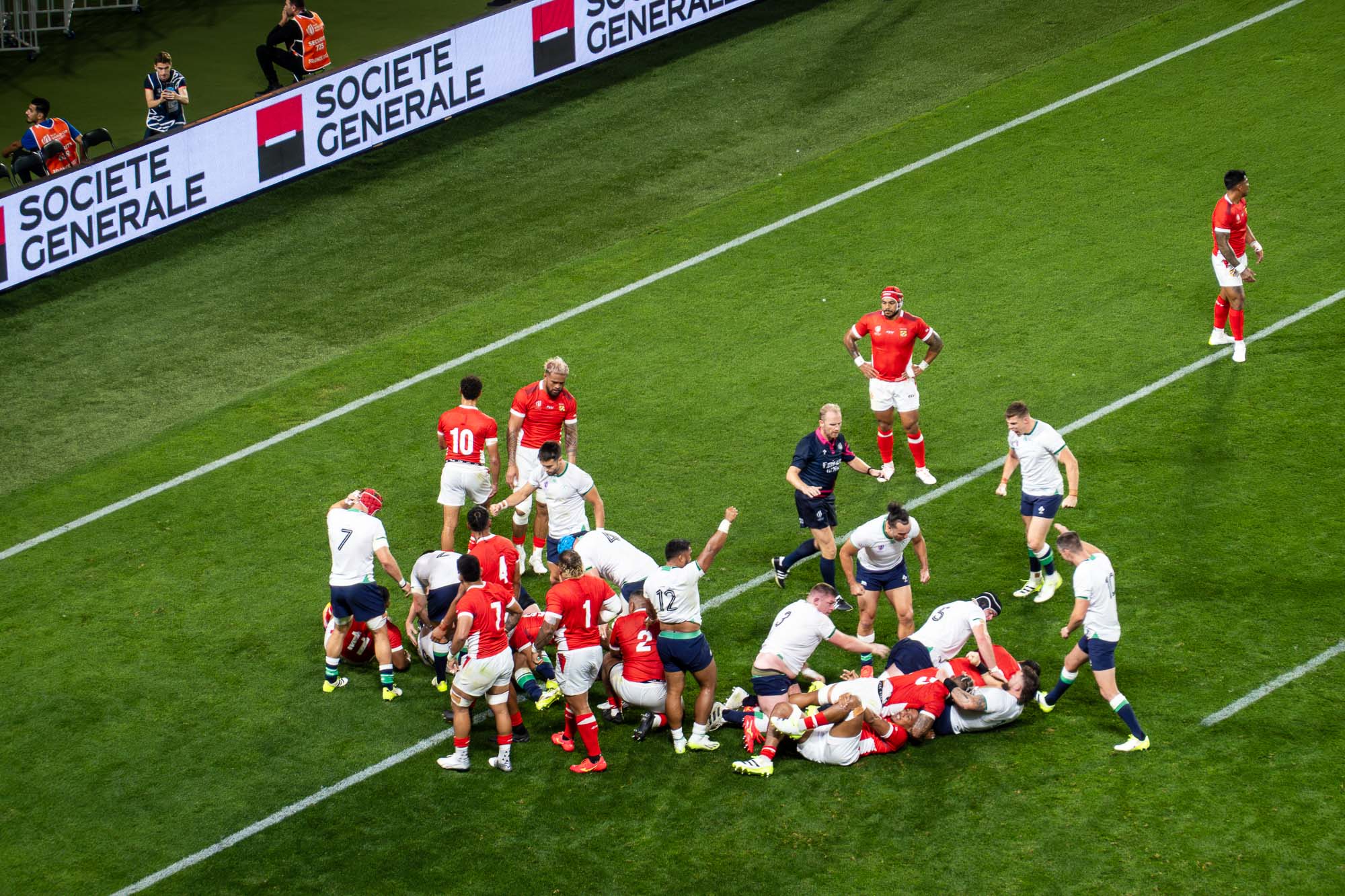 Regroupement lors d’Irlande – Tonga vu des tribunes : Tongiens en rouge, Irlandais en blanc, un joueur lève le poing, l’arbitre en bleu suit le jeu, panneaux LED Société Générale et stadier en gilet orange en haut à gauche