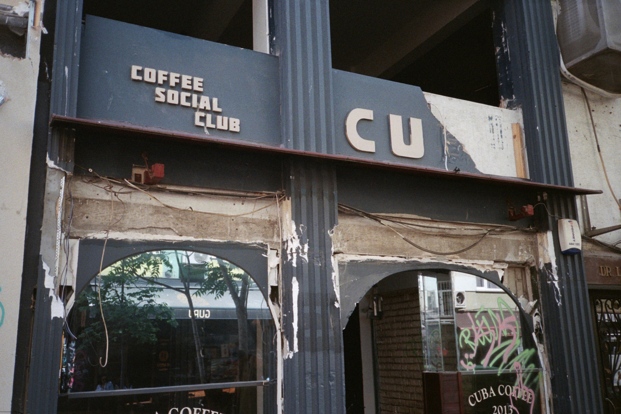 A weathered storefront of "Coffee Social Club" with partially missing signage, featuring arched windows and visible wiring against a city street backdrop.