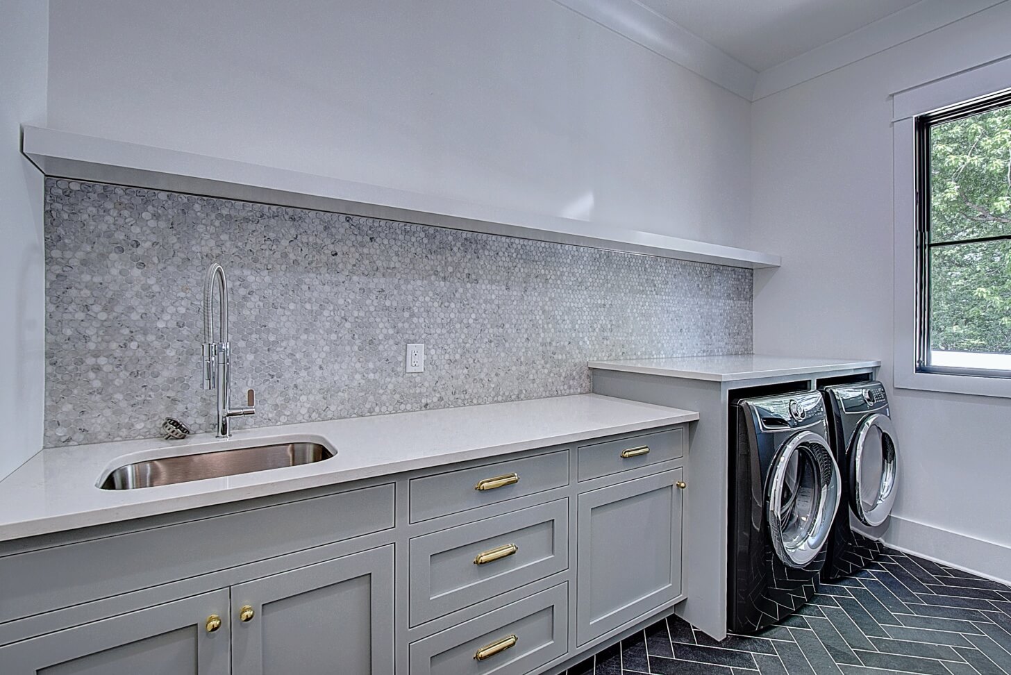Modern laundry room featuring a washing machine, gray cabinetry, and a marble backsplash. Natural light from a window.