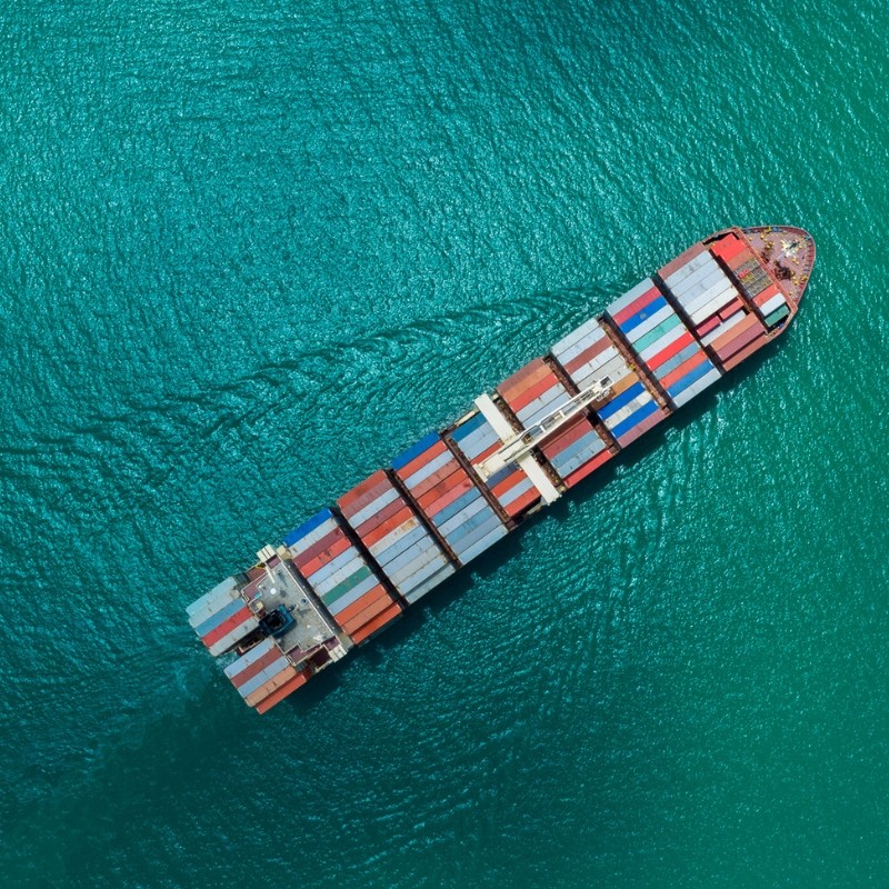 Overhead view of a large cargo ship loaded with colorful shipping containers sailing through turquoise water. A dark overlay features the heading “Import,” subtext “Saudi approvals & Arabic labels, end to end,” and a yellow button labeled “Learn More.”