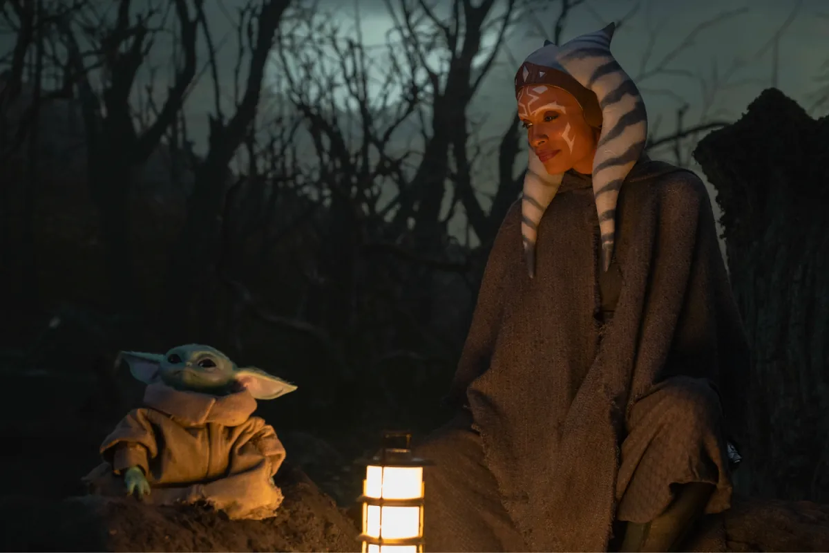 Grogu and Ahsoka Tano sit together in a dimly lit forest, illuminated by the soft glow of a lantern. Ahsoka smiles warmly as she observes Grogu, their bond forming as she uncovers his past and true name through the Force.