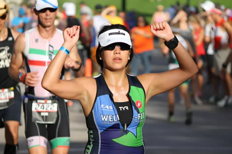 Female triathlete in Well-Fit race kit raising her fists in celebration during an Ironman run surrounded by spectators