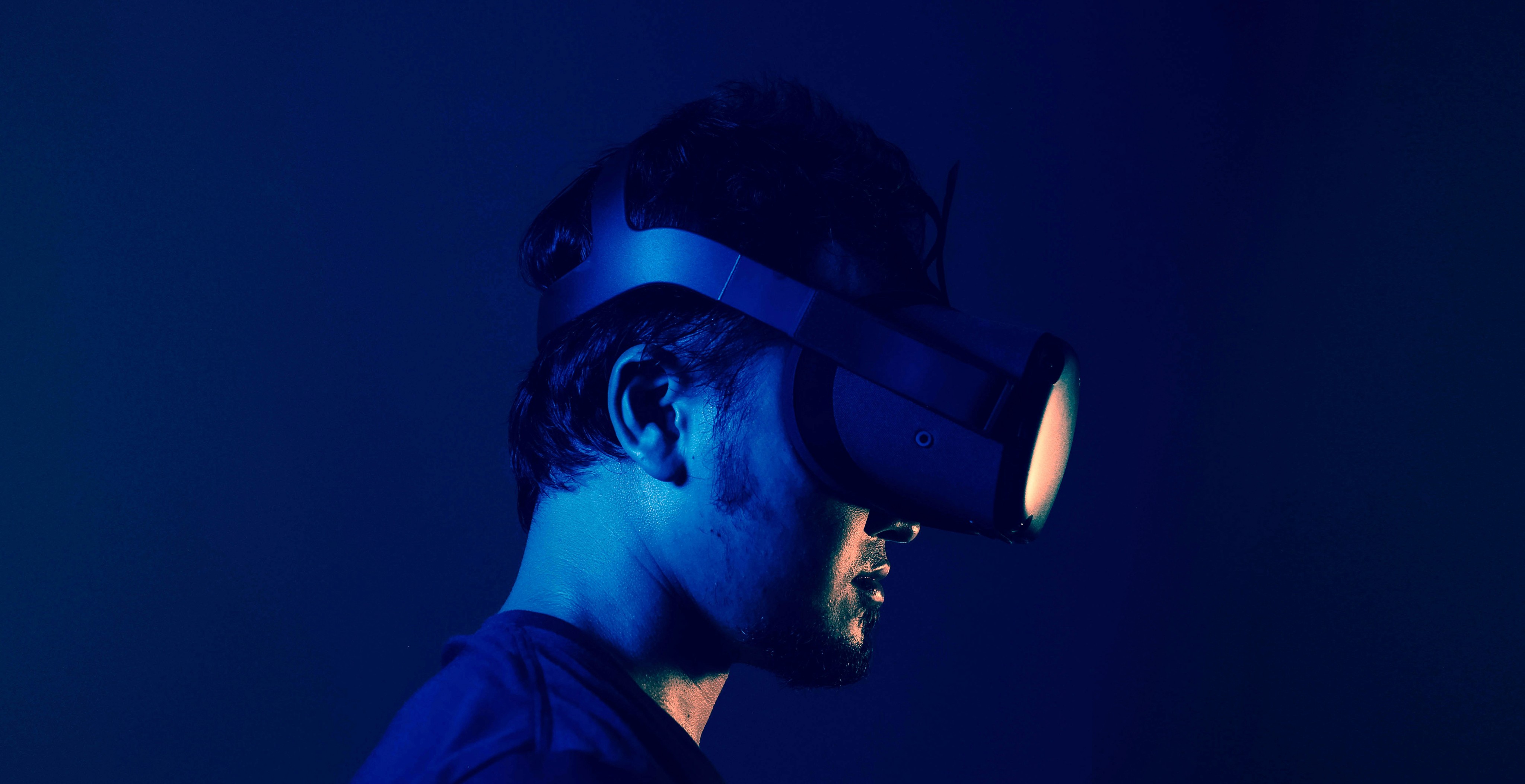 A person seen from the side profile wears a virtual reality (VR) headset in a dark room, illuminated by dramatic blue lighting and a warm glow coming from the headset's lens.