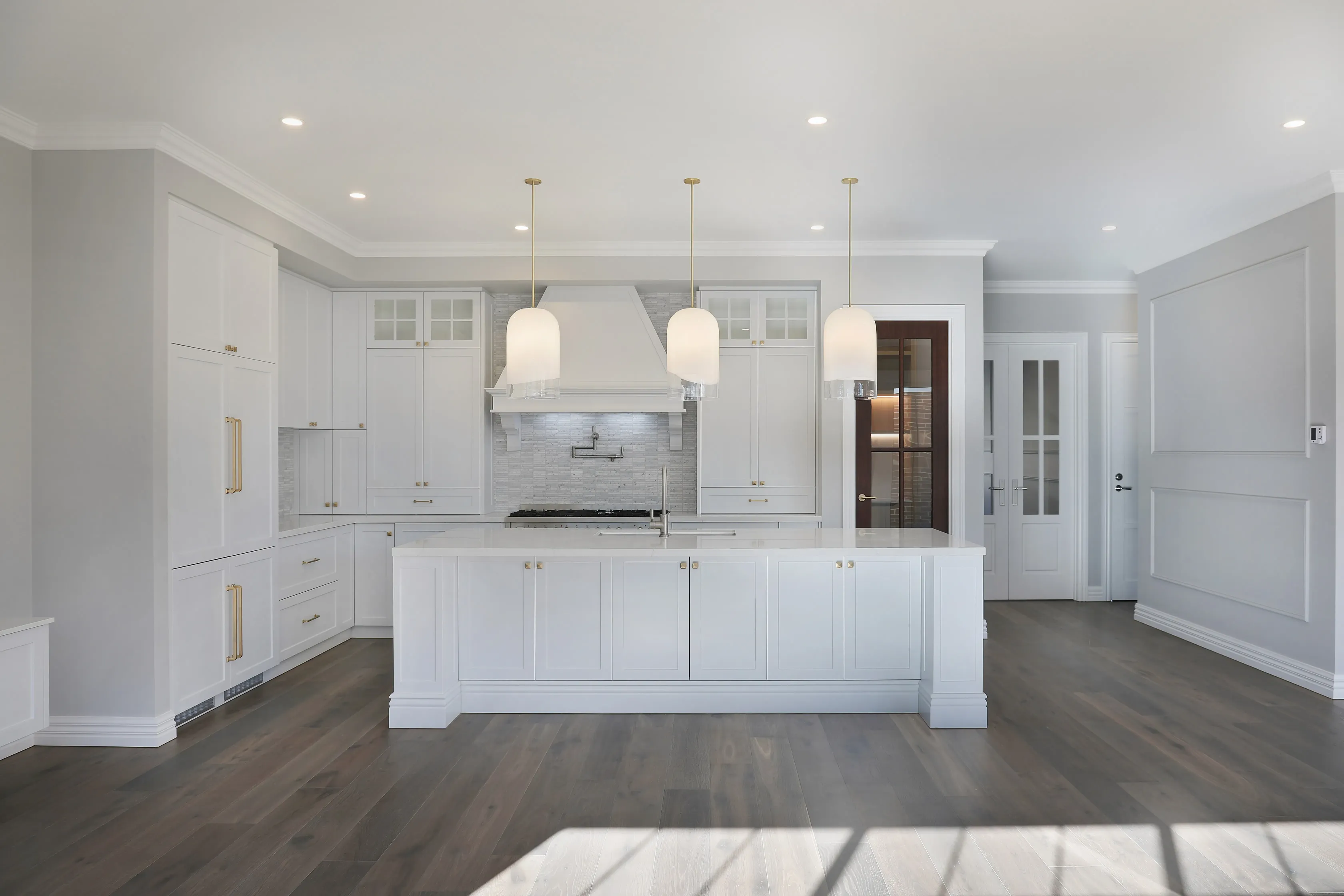Hamptons kitchen with white shaker cabinetry, brass hardware, three white dome pendants, a white range hood, marble mosaic splashback, a large white island and dark timber floors