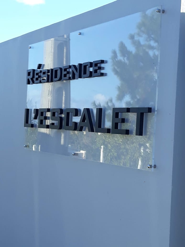 Panneau indiquant « Résidence L'Escalet » fixé sur un mur blanc. Le panneau transparent reflète les arbres et le ciel, suggérant un environnement serein et haut de gamme.
