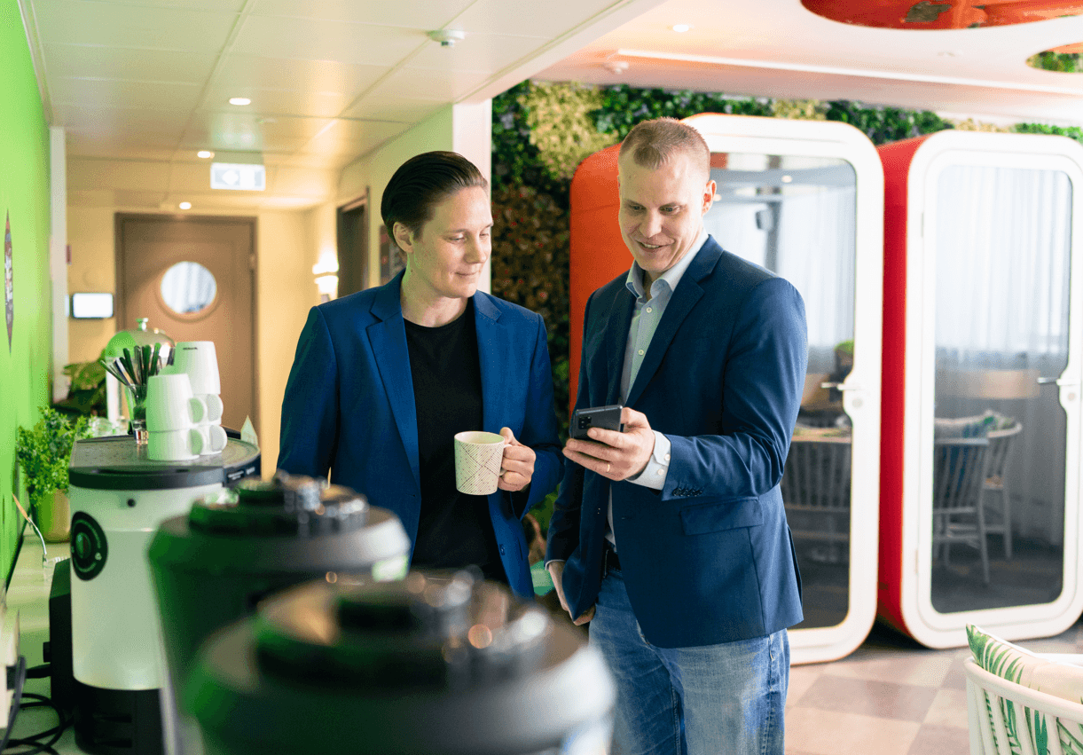 Tuulia Virhiä and Juhani Heinonen in business attire stand in an office, one holding a coffee mug, the other looking at a smartphone. They appear to be in discussion