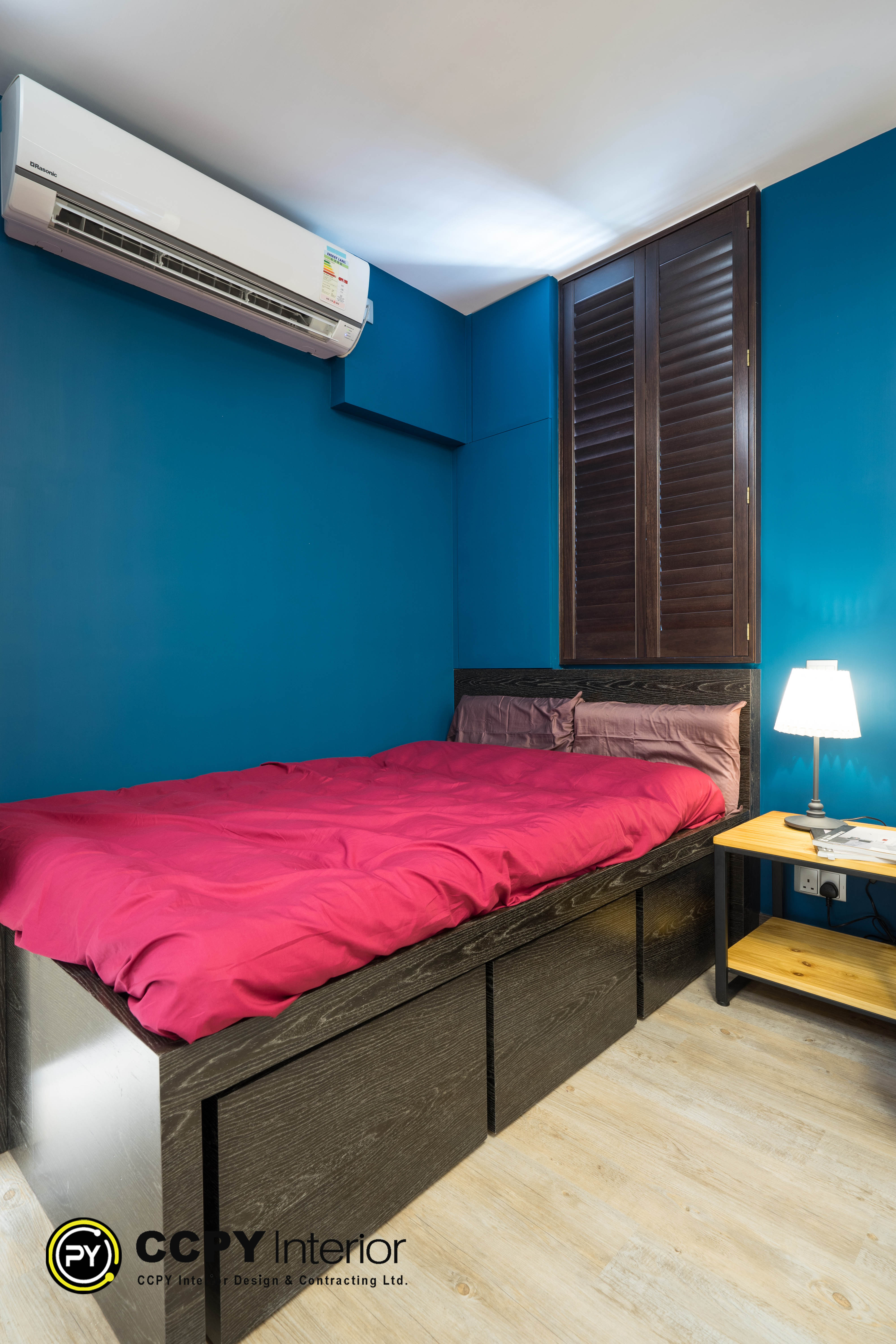 The selected region displays an interior bedroom design featuring a bold color scheme and functional layout.Visual FeaturesWalls: Painted in a deep, matte royal blue color.Bedding: Fitted with a vibrant magenta or deep pink duvet cover and matching pillows.Furniture: A dark wood headboard paired with a light wood bedside table.Lighting: A warm-toned, modern bedside table lamp illuminating the corner.Window: Fitted with dark, horizontal wooden shutters or blinds.