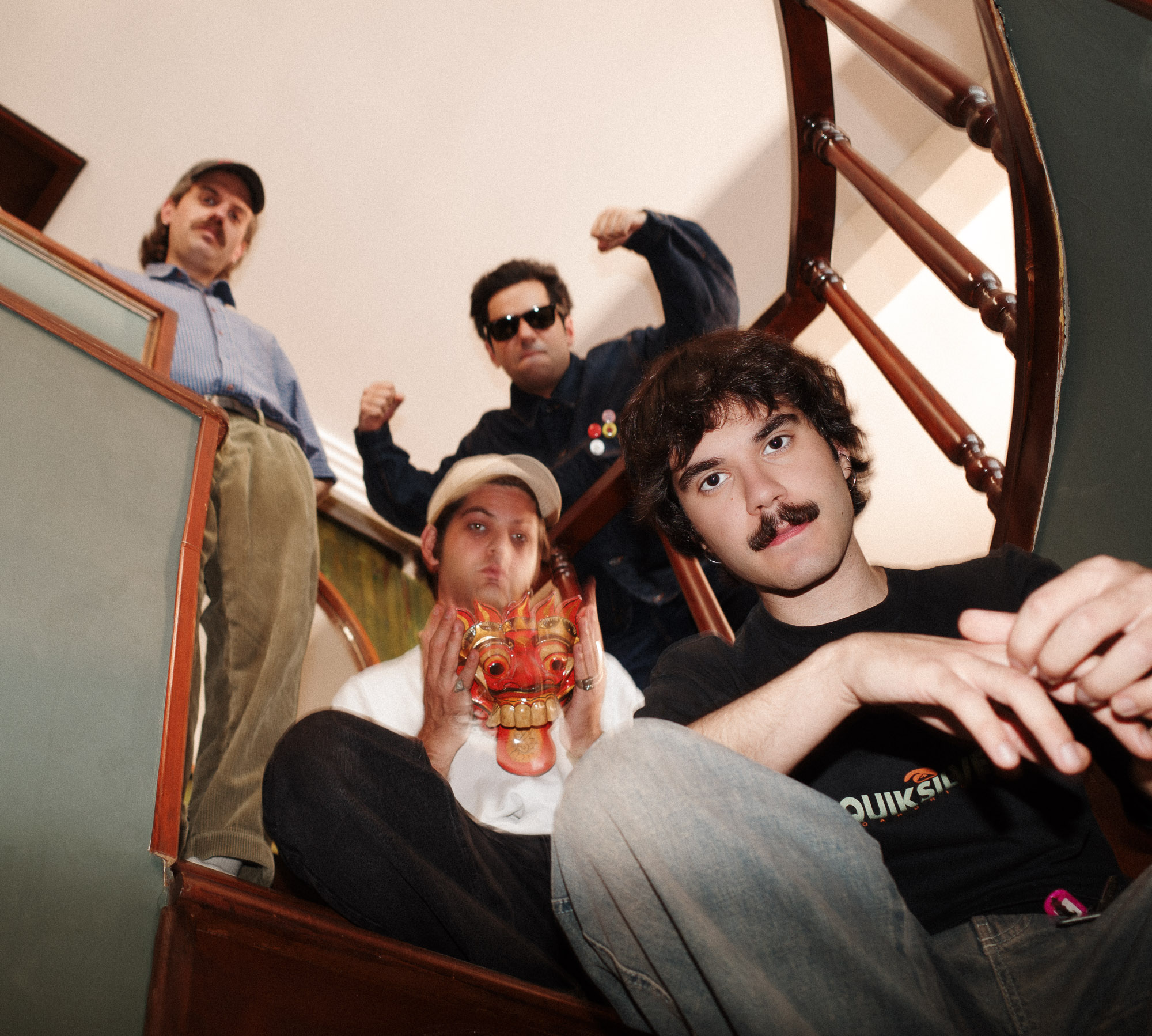 A group of four casually dressed people posing on a staircase, one holding a colorful dragon mask, with wooden railings in the background, conveying a relaxed and playful atmosphere.