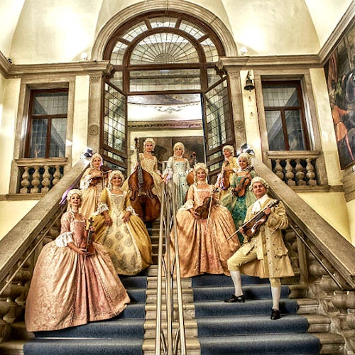 Gruppo di persone in elaborati costumi d'epoca, con strumenti musicali in mano, in piedi sulla grande scalinata di un edificio lussuoso.