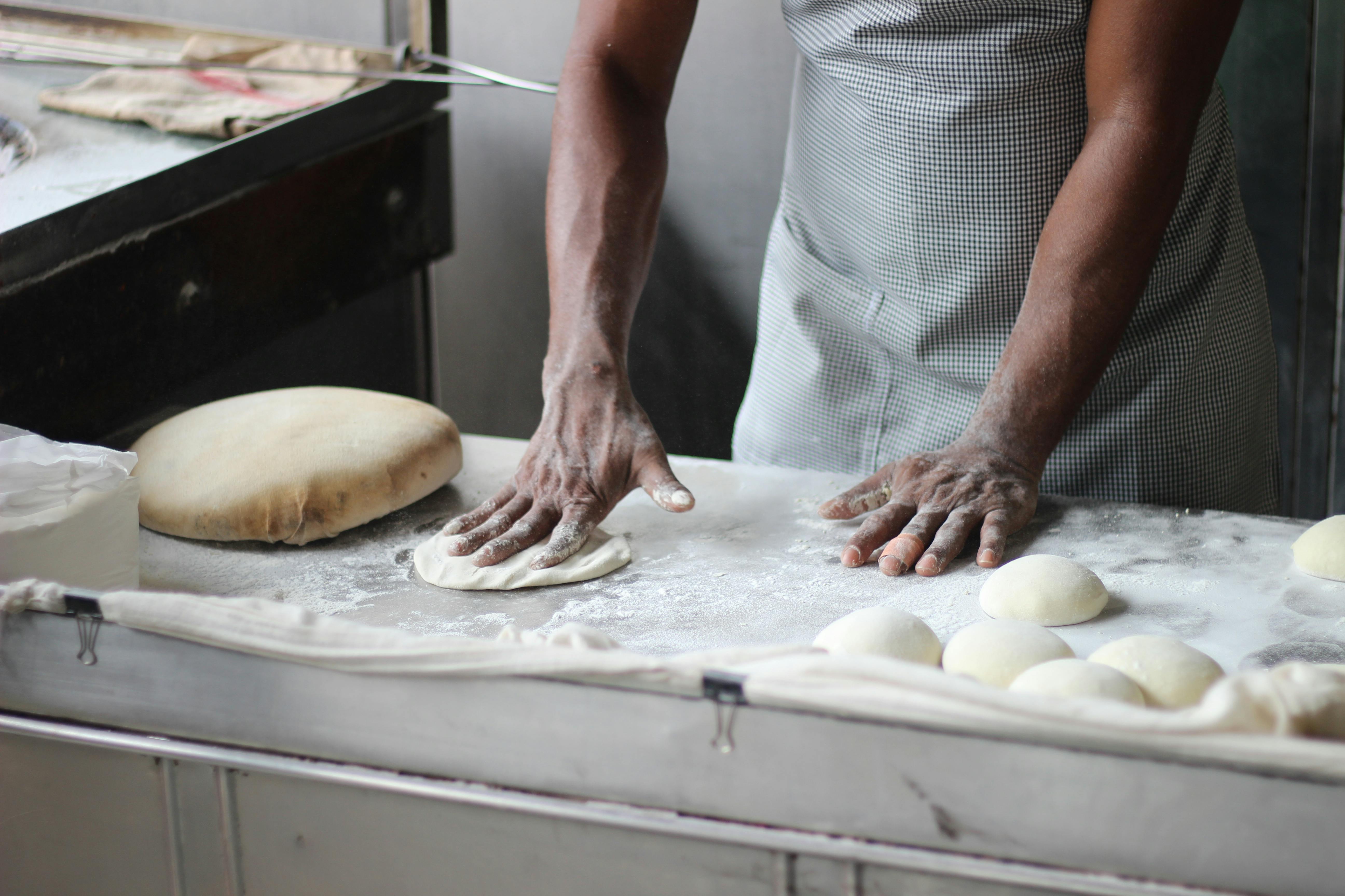 Dettaglio professionale di un uomo che impasta a mano un panetto di pane su un piano di lavoro, con altri panetti già formati disposti in ordine sul piano