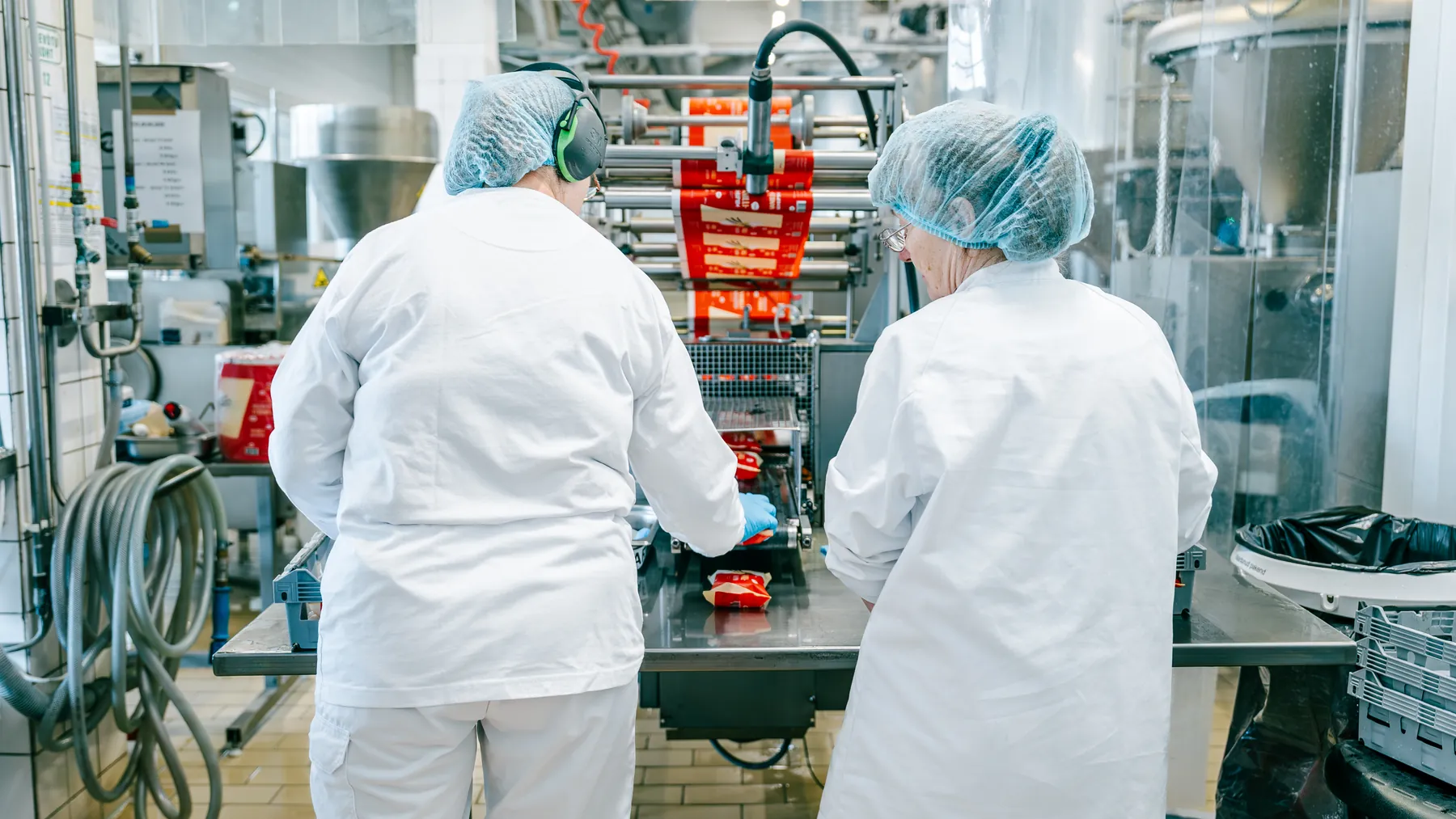 Two workers in hairnets and ear protection monitoring a packaging machine