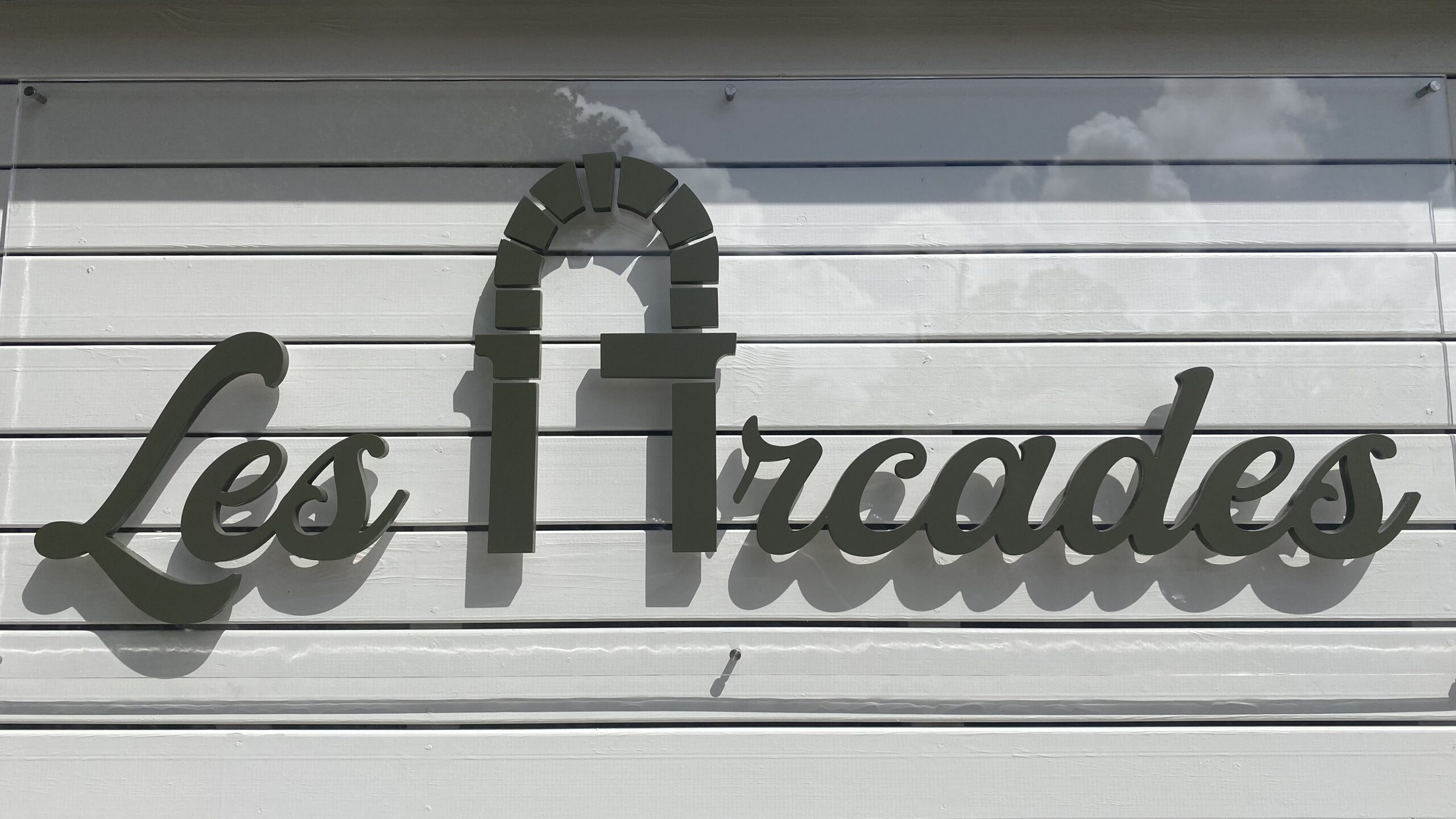 Enseigne Exterieur portant l'inscription « Les Arcades » en cursive, avec une arche intégrée. Elle est fixée sur des panneaux blancs sous un ciel nuageux, créant une ambiance accueillante.