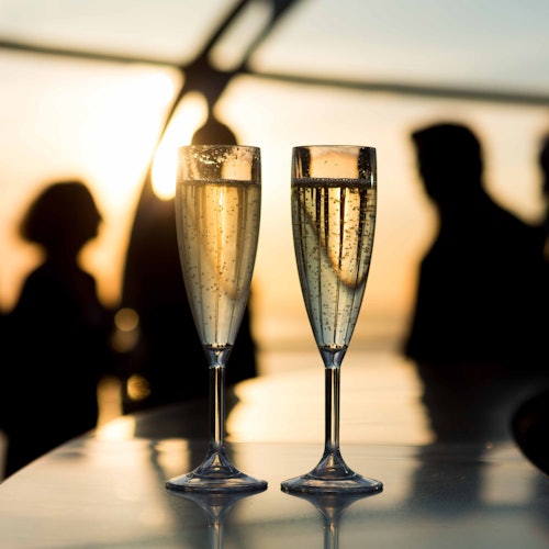 Two champagne flutes on a table with sunset-lit silhouettes of people in the background.