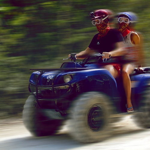 Duas pessoas usando capacetes, óculos de proteção e bandanas pilotam um quadriciclo azul em alta velocidade por uma área arborizada e embaçada.