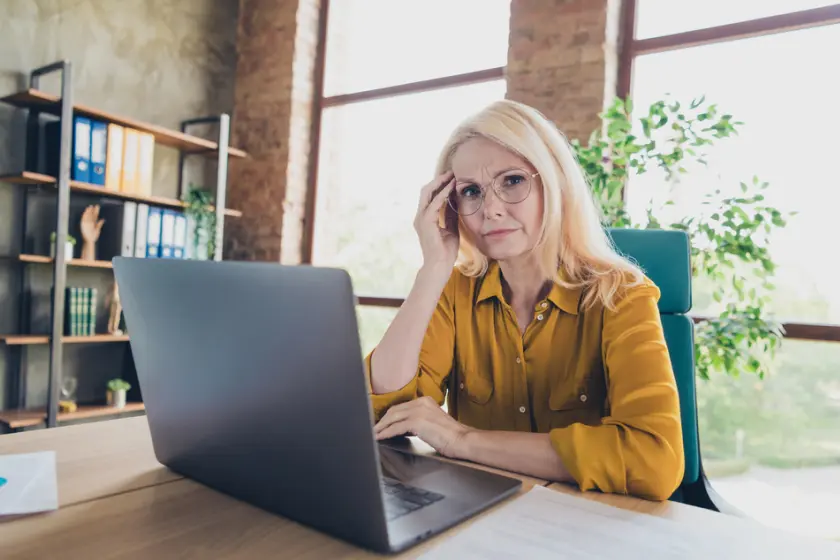 Mature woman wearing glasses, sitting at a desk with a laptop, looking thoughtful in a modern office setting.