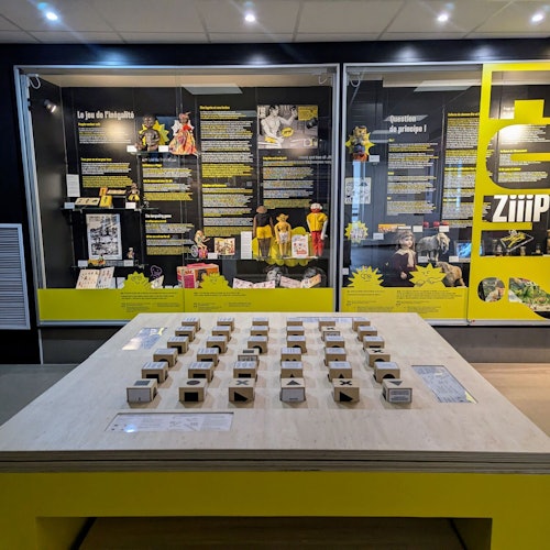 Utställning med textpaneler, gula accenter och interaktiva spelbitar på ett bord, allt inramat inom ett visningsrum.