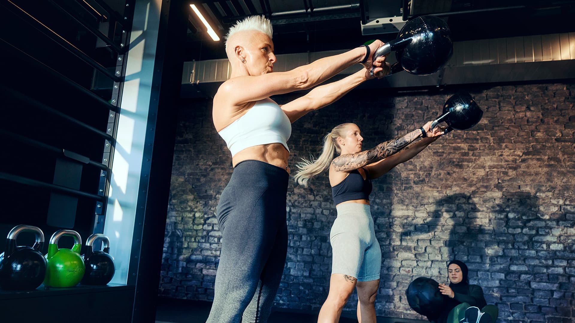 Zwei Frauen trainieren intensiv mit Kettlebells in einem modernen Fitnessstudio, während eine dritte Person mit einem Medizinball im Hintergrund sitzt.