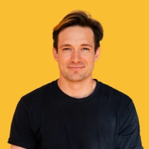 A person with short brown hair wearing a black t-shirt stands smiling in front of a vibrant yellow background.
