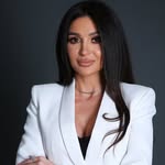 Young person with wavy hair poses confidently in sunlight against a modern building backdrop. Wears a black blazer and silver necklace, conveying elegance.