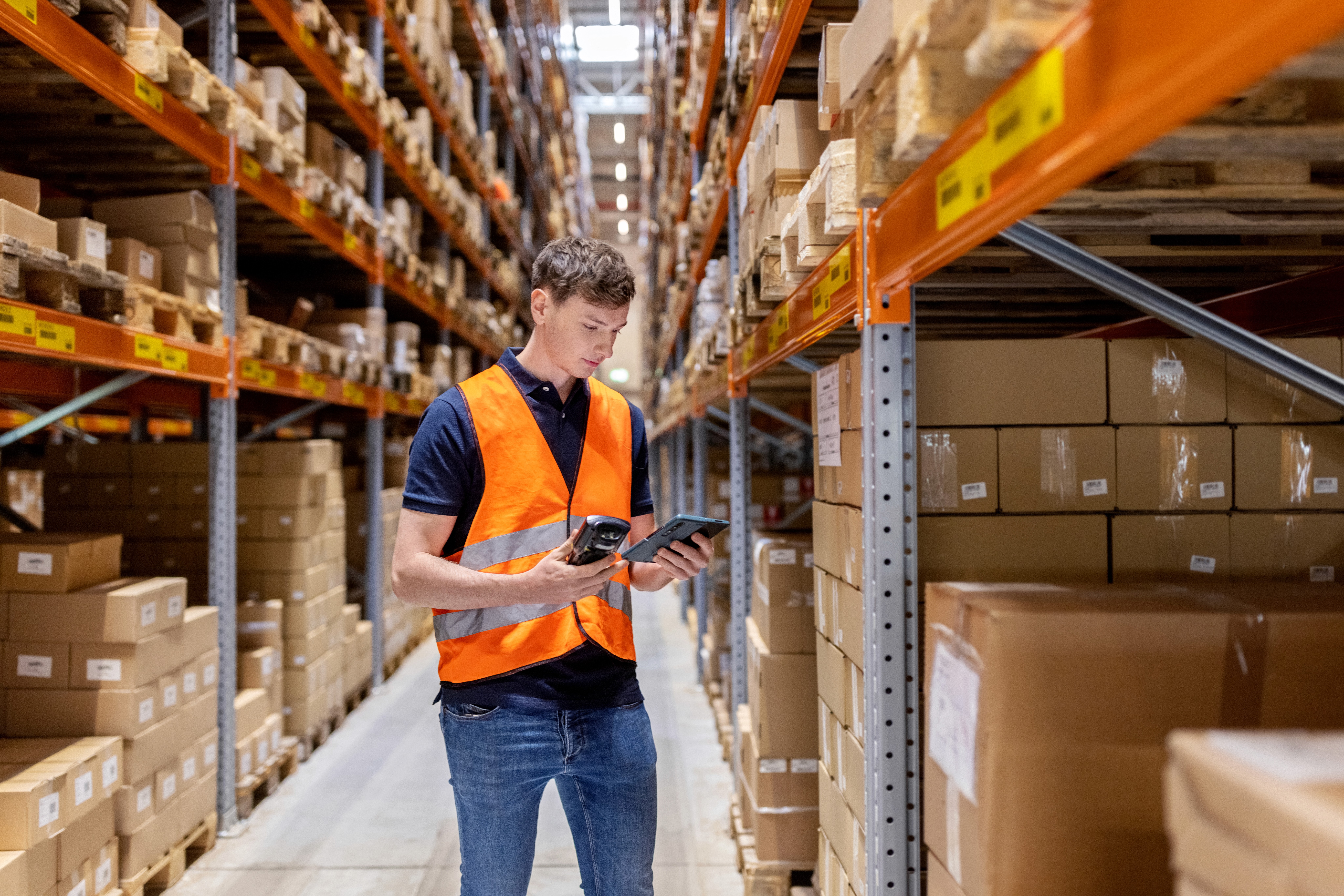 A warehouse worker wearing an orange safety vest is holding a tablet, standing in a spacious aisle lined with tall shelves filled with neatly stacked cardboard boxes.