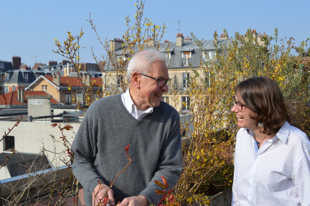 Un homme âgé et une auxiliaire de vie rient ensemble en taillant des arbustes sur une terrasse parisienne
