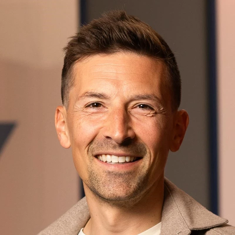 A person with short, styled hair wears a light jacket over a white shirt. The background features a neutral indoor wall with subtle geometric shapes.
