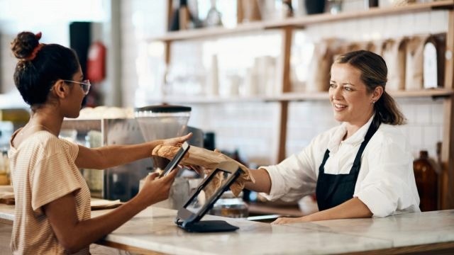 Em ambiente de padaria, mulher branca entrega item a mulher de pele parda e cabelo amarrado em coque, do outro lado do balcão.
